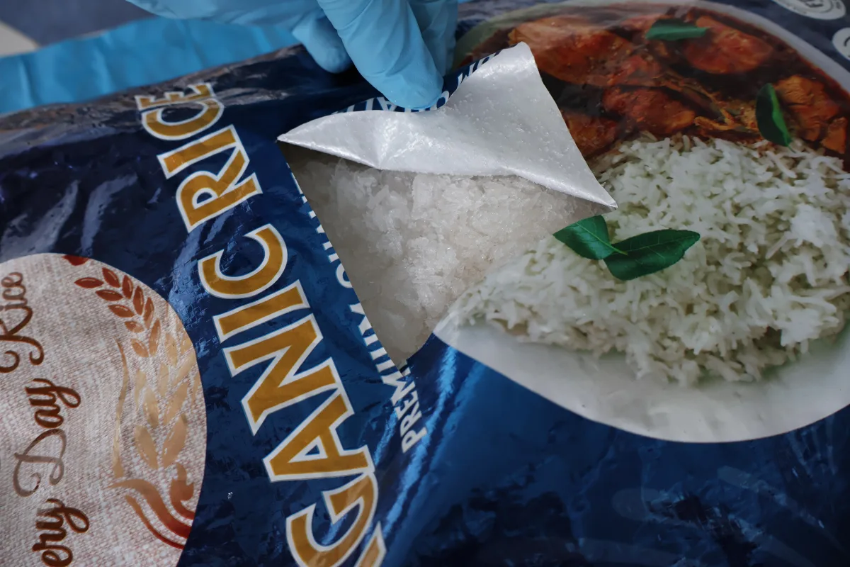 Eine Hand mit blauem Handschuh öffnet eine Reistüte, um eine unbekannte weiße Substanz zu zeigen. Die Tüte trägt das Etikett 'Organic Rice'.