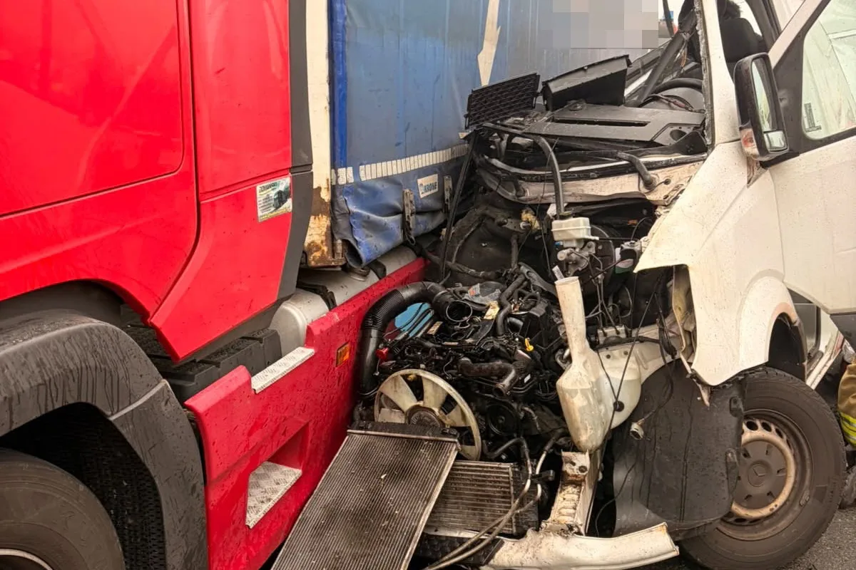Das Bild zeigt einen stark beschädigten weißen Lieferwagen, der frontal mit einem großen roten LKW zusammengestoßen ist. Der Motorraum und die Front des Lieferwagens sind stark eingedrückt und es sind Motorenteile sichtbar.