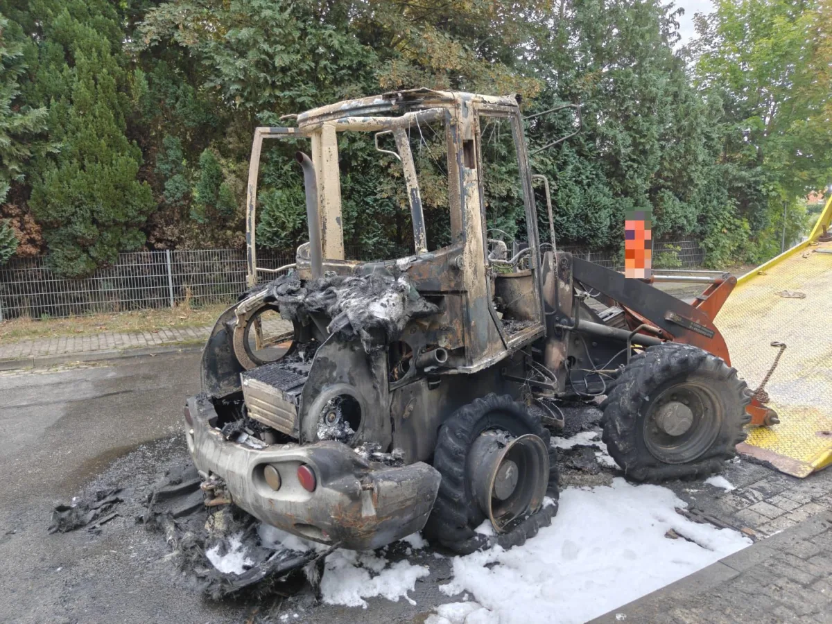 Ein ausgebrannter Bagger steht auf einer Straße, teilweise von Löschschaum bedeckt. Dichte Vegetation ist im Hintergrund sichtbar.