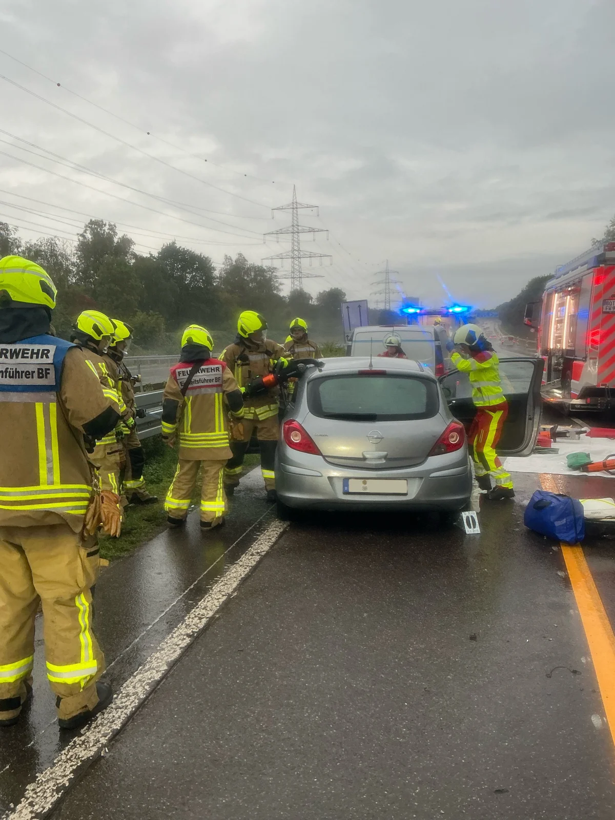 Feuerwehrleute in gelben Helmen und Schutzkleidung stehen um ein silberfarbenes Auto auf einer regennassen Straße. Ein Rettungsfahrzeug mit blinkenden Blaulichtern ist im Hintergrund sichtbar.