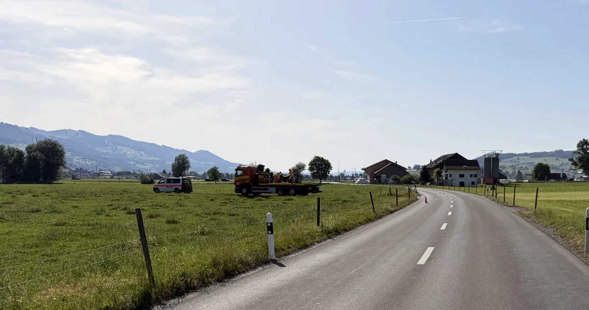 Ein Abschleppwagen steht auf einem Feld neben einer Landstraße, begleitet von einem Polizeiauto. Die Landschaft ist von Hügeln und vereinzelten Gebäuden geprägt.