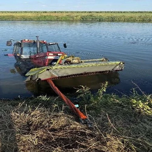 Ein roter Traktor mit angeschlossenem Mähwerk steckt halb im Wasser eines Grabens am Feldrand fest. Im Hintergrund sind Felder und blauer Himmel zu sehen.