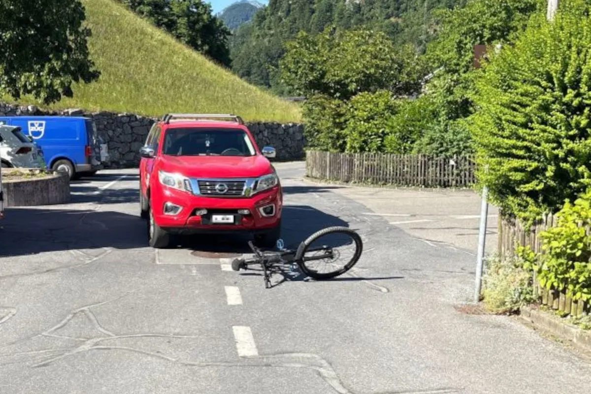 Auf einer Straße liegt ein Fahrrad vor einem roten Auto. Im Hintergrund sind Bäume und ein Hügel sichtbar.