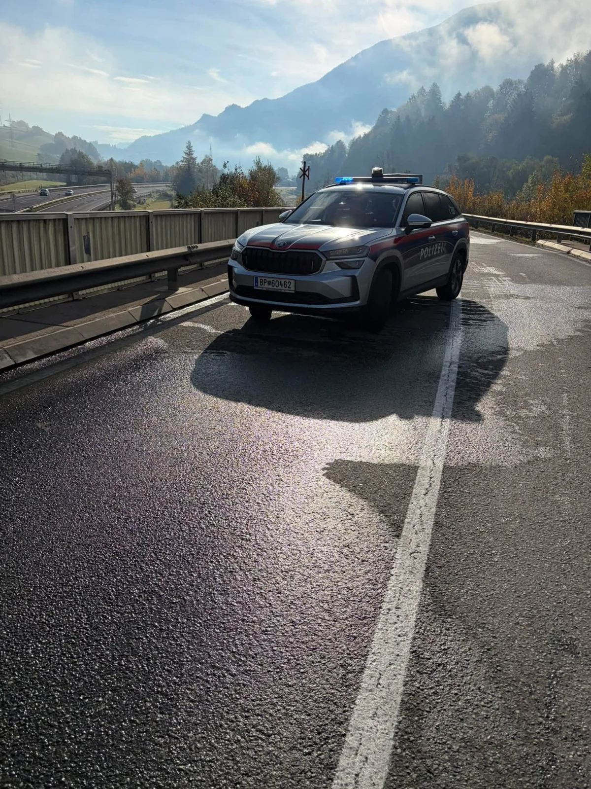 Ein Polizeifahrzeug steht auf einer Landstraße. Im Hintergrund sind nebelbedeckte Hügel und dichte Bewaldung zu sehen.