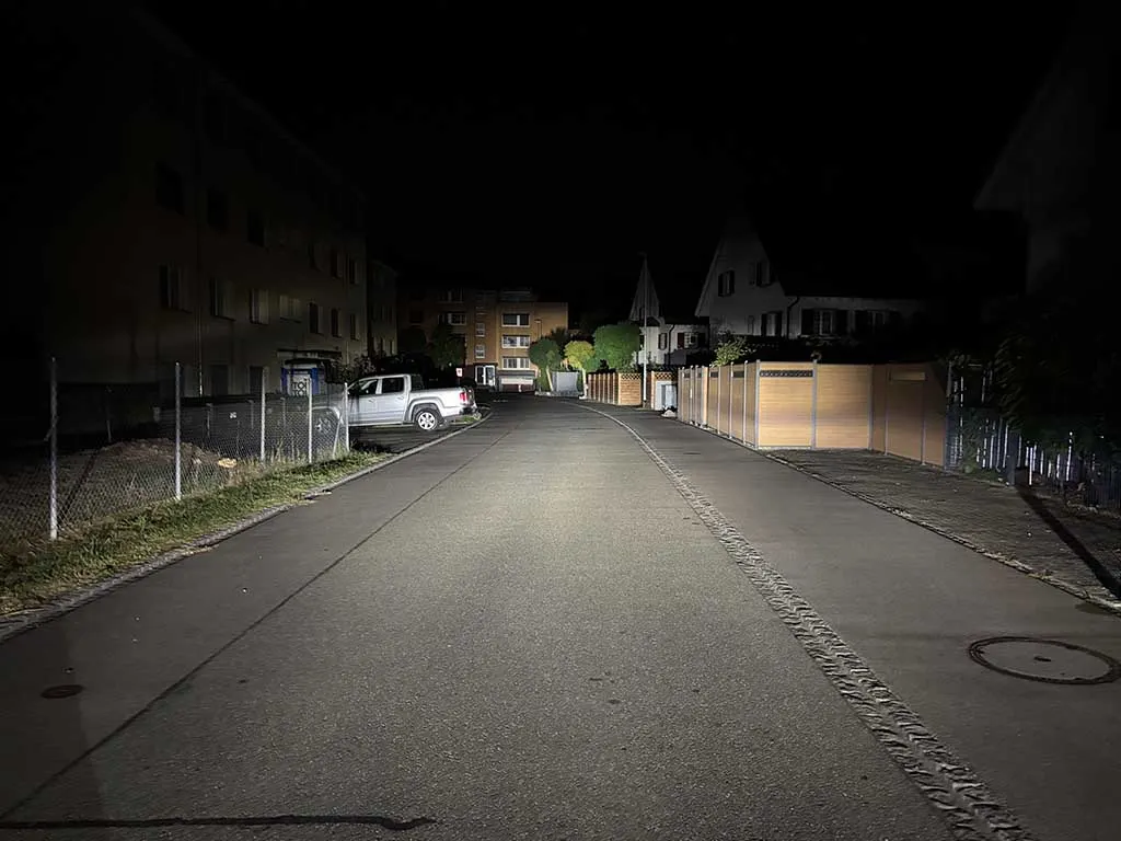 Nachtaufnahme einer Wohnstraße, beleuchtet von einem Fahrzeug. Links ein geparktes Auto, rechts Wohnhäuser.