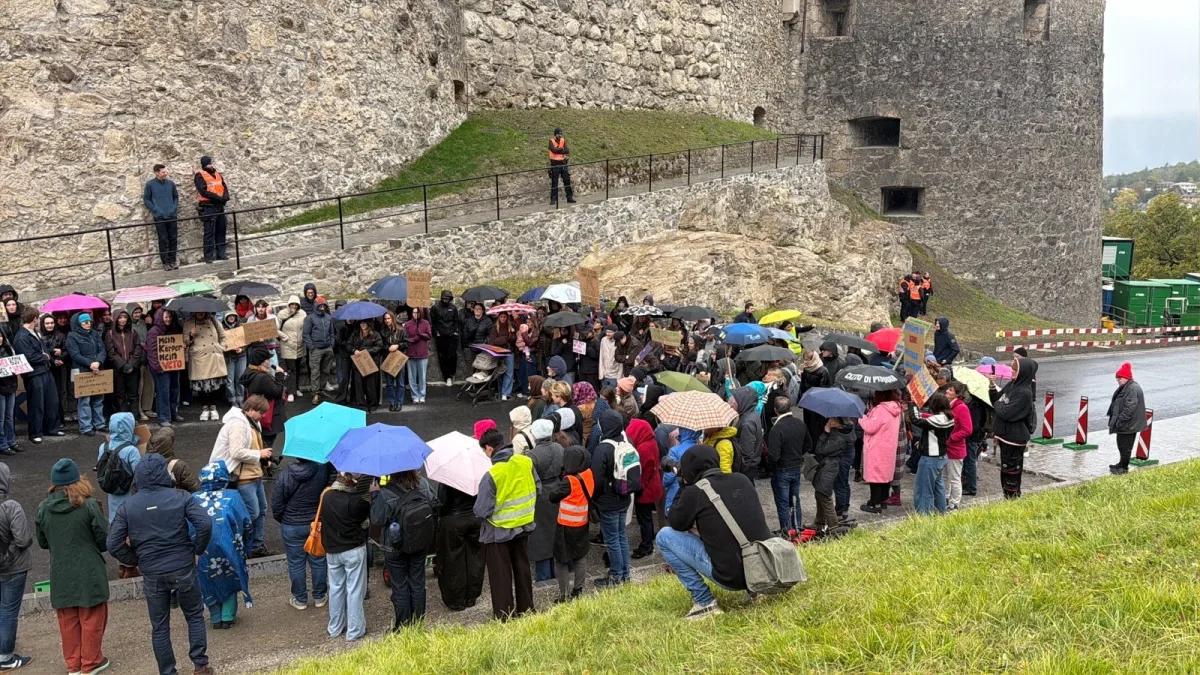 Eine große Gruppe von Menschen steht mit Regenschirmen vor einer alten Steinmauer. Einige tragen Schilder, während mehrere Polizisten in orangenen Westen die Menge beobachten.