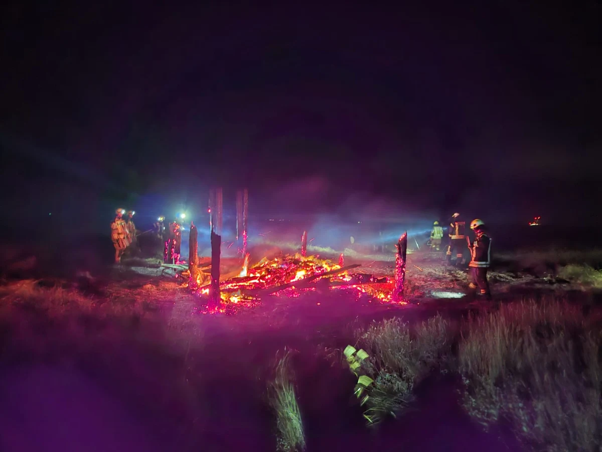 Mehrere Feuerwehrleute in Schutzausrüstung löschen einen Brand auf einem dunklen Feld. Glühende Überreste sind sichtbar.