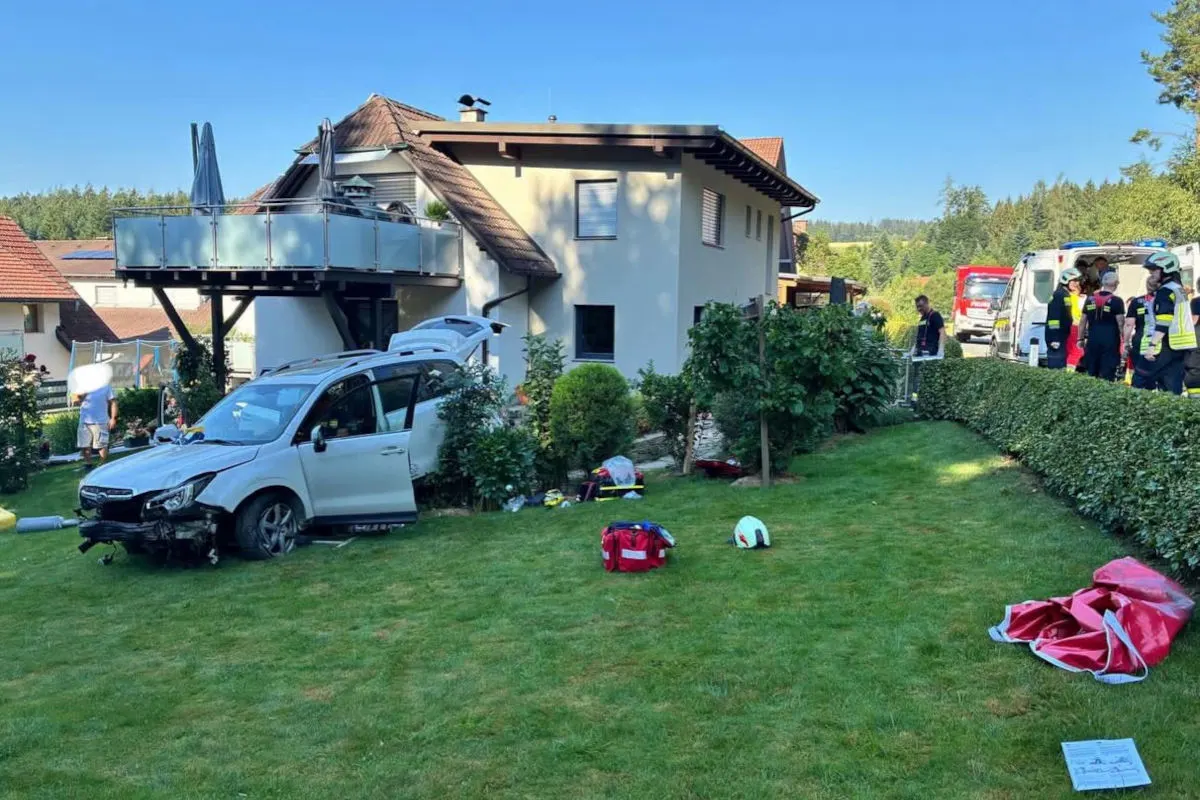 Ein beschädigtes Auto steht im Garten eines Hauses. Rettungskräfte sind im Hintergrund zu sehen.