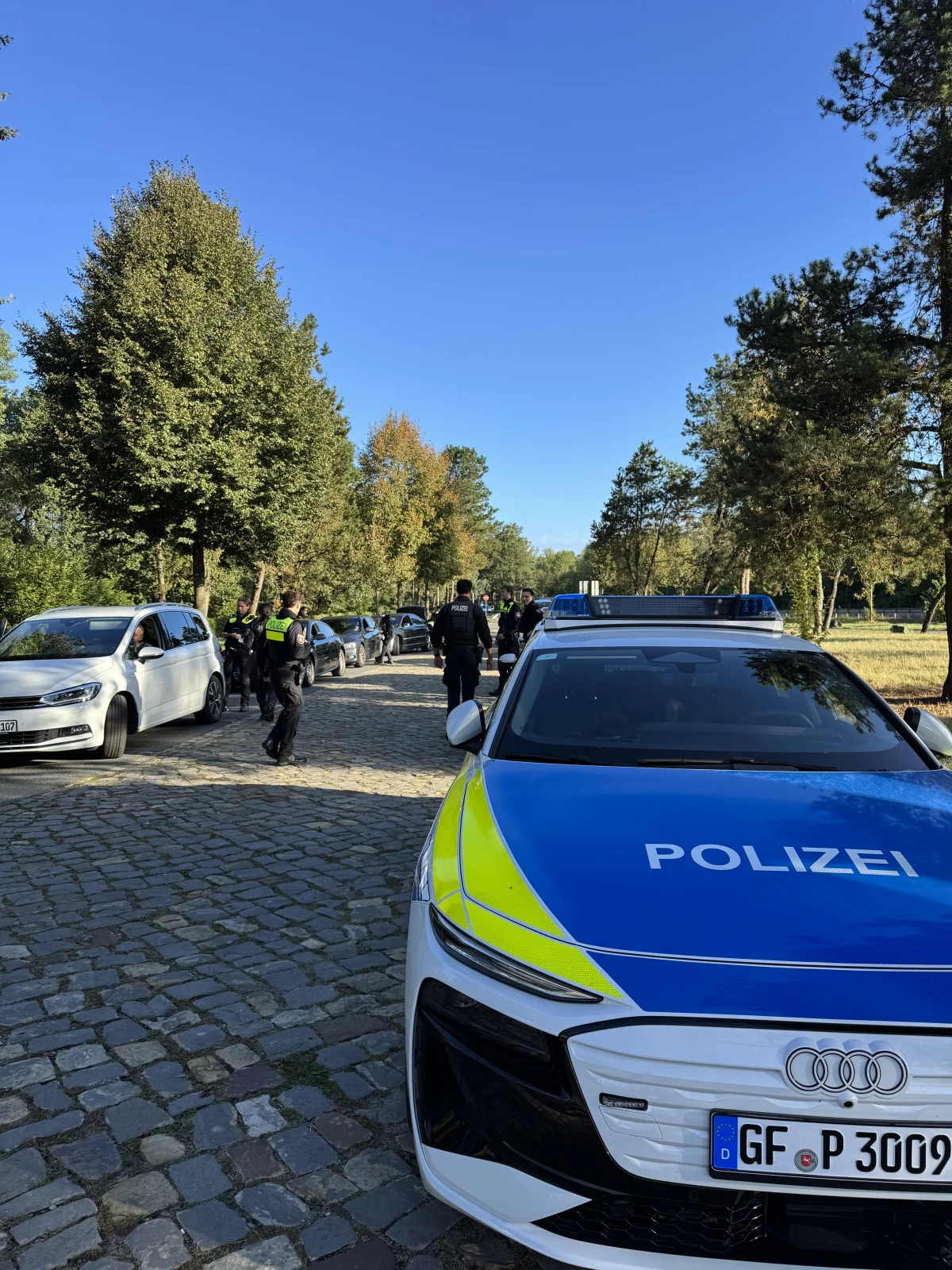 Ein Polizeifahrzeug mit der Aufschrift 'Polizei' steht auf einer Kopfsteinpflasterstraße, umgeben von Beamten und Fahrzeugen. Es ist Tag und der Himmel ist klar.