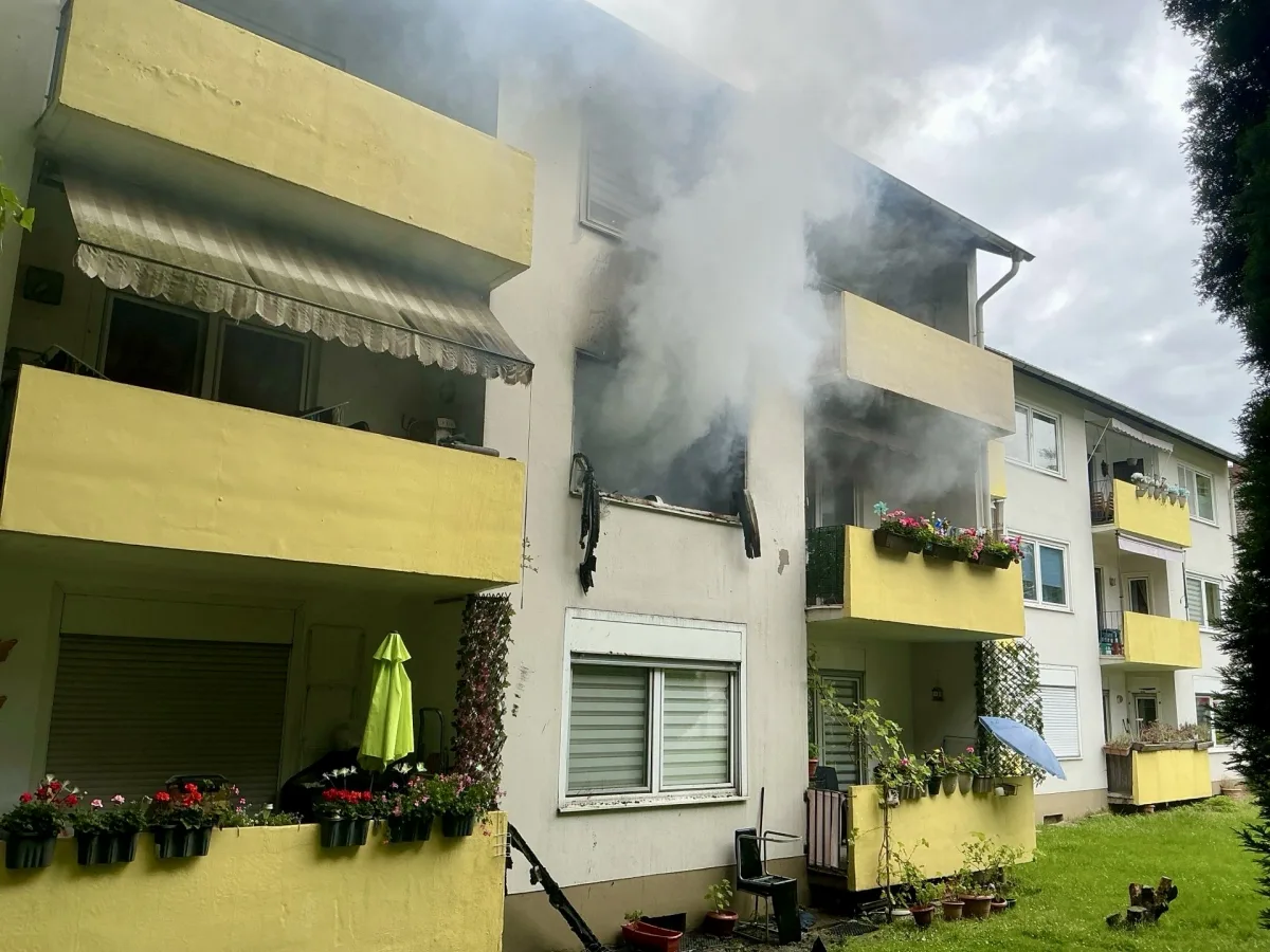 Dichtes Rauchaufkommen tritt aus einem Balkon im zweiten Stock eines mehrstöckigen Wohnhauses aus, während Pflanzenkästen an den Balustraden sichtbar sind.
