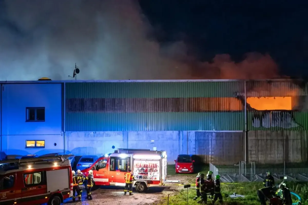 Ein großes, grünes Gebäude brennt; Rauch steigt auf, während Feuerwehrfahrzeuge und Einsatzkräfte gut sichtbar davorstehen.