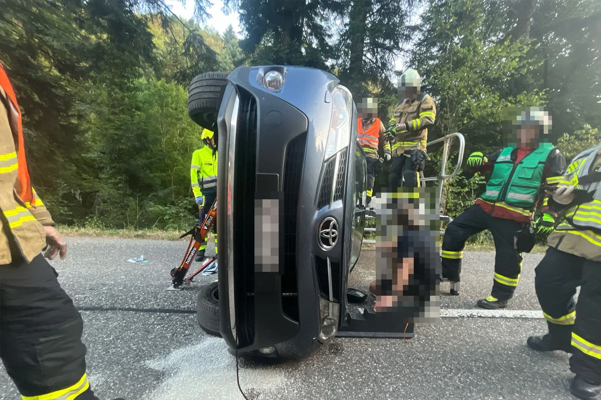 Ein Auto liegt auf der Seite auf der Straße, während mehrere Rettungskräfte in Sicherheitskleidung um das Fahrzeug herum stehen.