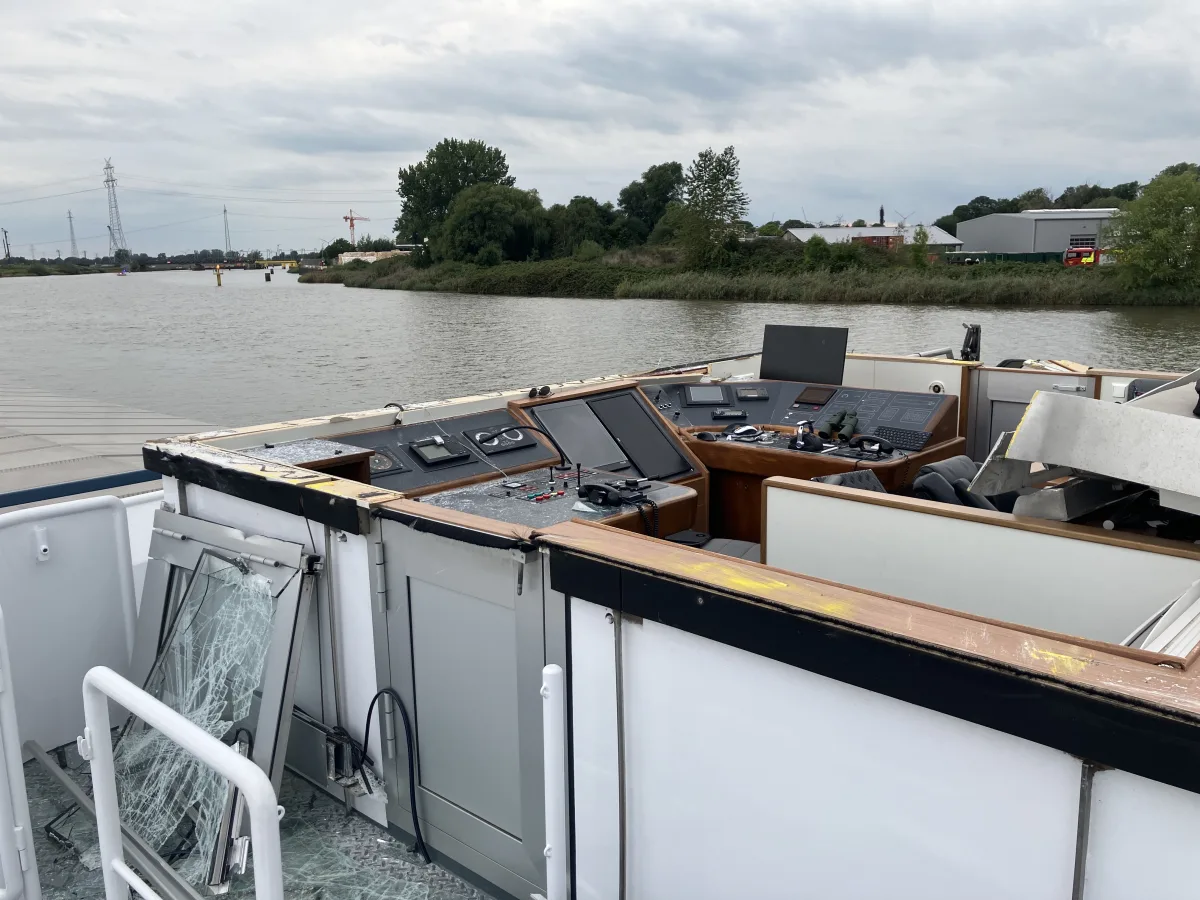 Das Bild zeigt ein Steuerhaus auf einem Schiff, das sichtbare Schäden aufweist, einschließlich zerbrochener Fenster. Im Hintergrund ist ein Fluss und Uferbewuchs zu sehen.