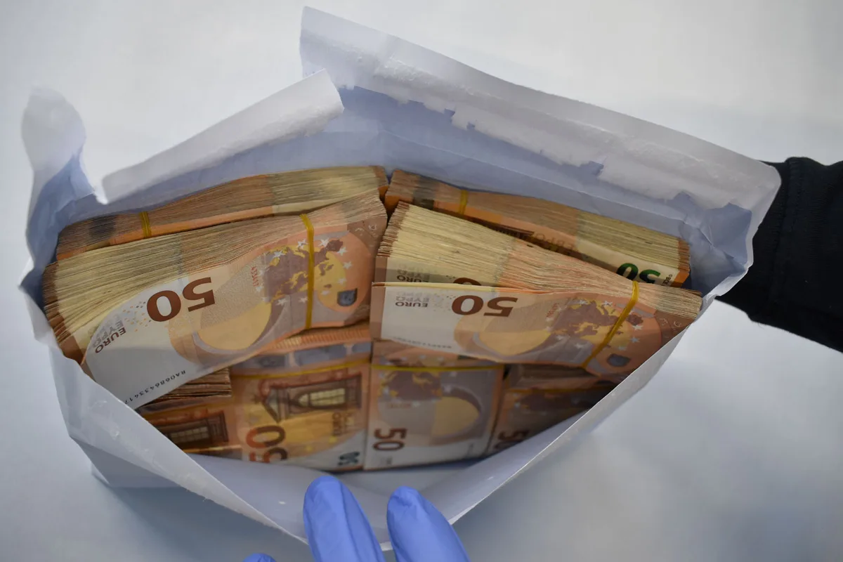 Ein geöffneter Umschlag, gefüllt mit Bündeln von 50-Euro-Scheinen, wird von einer Person mit blauen Handschuhen festgehalten. Die Scheine sind sorgfältig mit Gummibändern gebündelt.