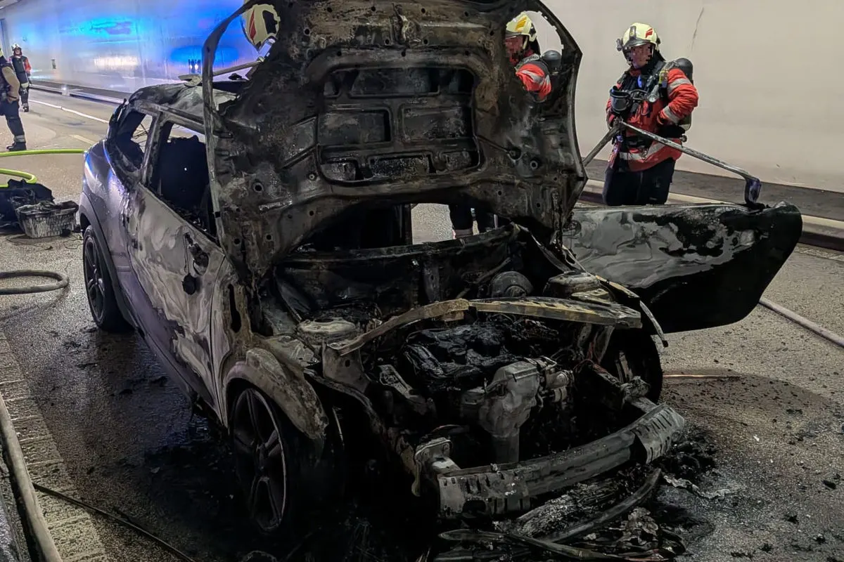 Ein stark verbranntes Auto steht in einem Tunnel. Im Hintergrund sind Feuerwehrleute zu sehen, die bei den Löscharbeiten sind.