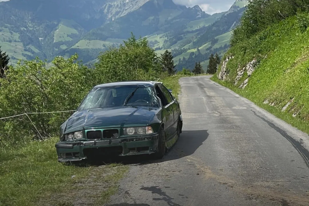 Ein grüner BMW mit sichtbaren Schäden steht am Rand einer schmalen, asphaltierten Bergstraße. Im Hintergrund sind grüne Hügel und Berge zu sehen.