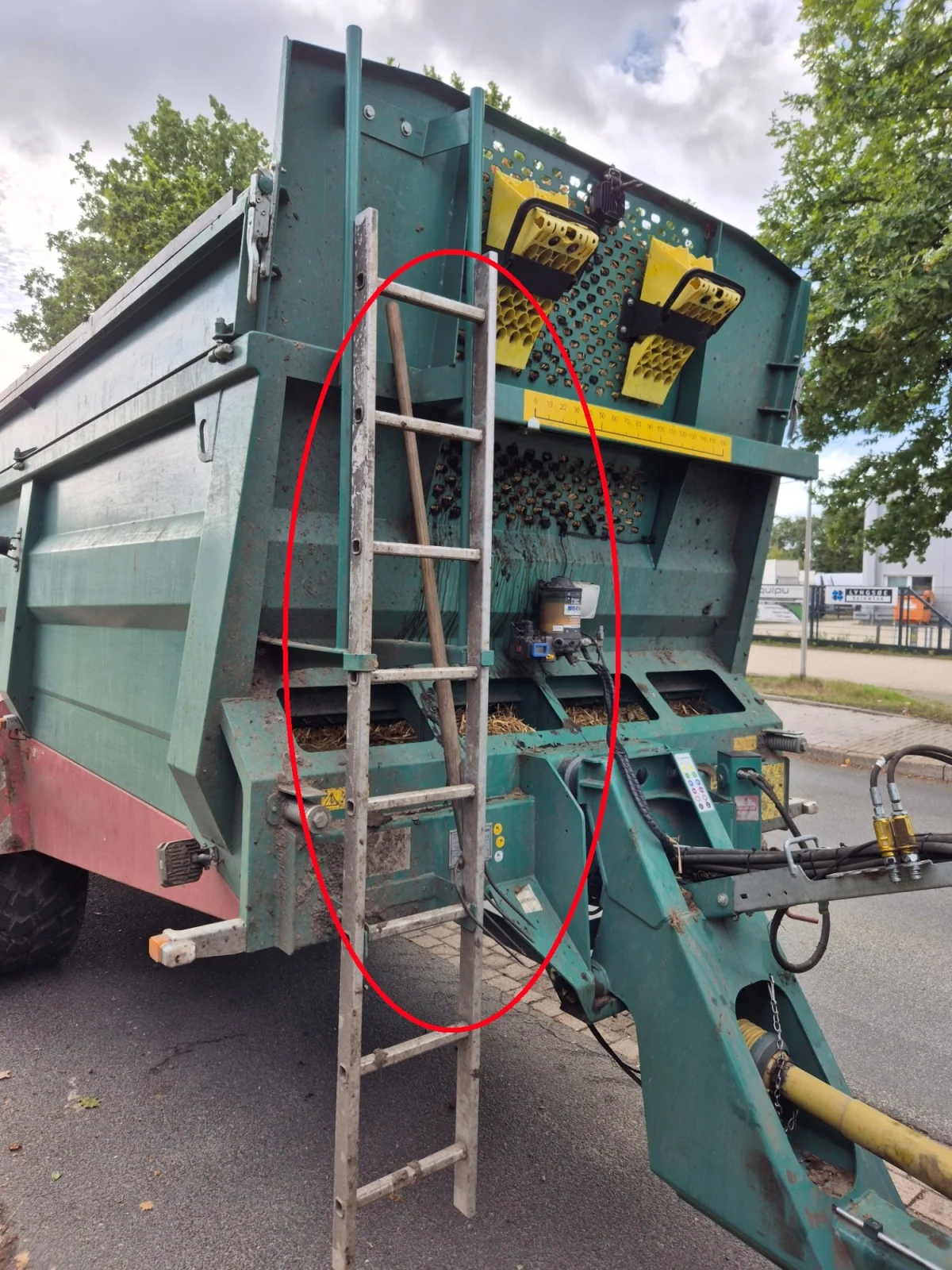 Ein grüner landwirtschaftlicher Anhänger steht an einer Straße. Eine Leiter ist an seiner Seite befestigt. Im Hintergrund sind Bäume und ein Gehweg sichtbar.