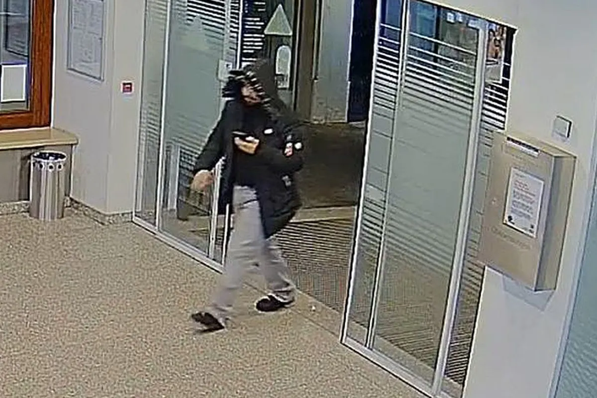 Eine Person in dunkler Kleidung mit einer Kapuze über dem Kopf betritt durch automatische Glastüren einen Innenbereich. Sie schaut auf ein Mobiltelefon in ihrer rechten Hand.