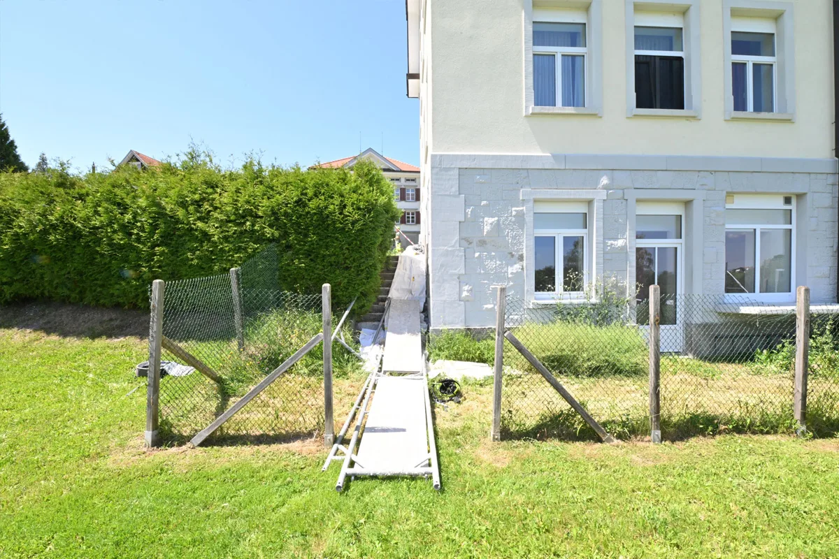 Eine provisorische Rampe erstreckt sich von einem Gartenzaun zu einem gelben Gebäude mit mehreren Fenstern.