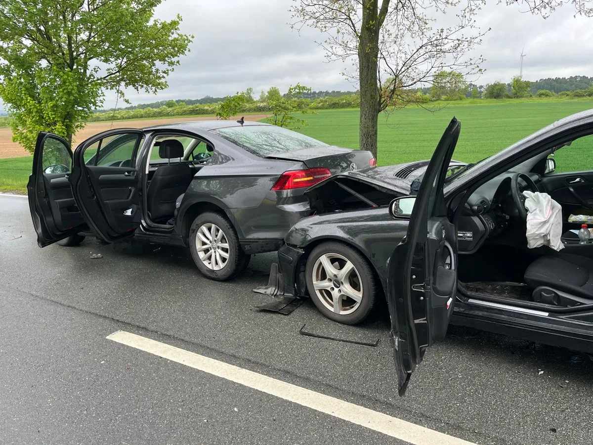 Zwei graue Fahrzeuge stehen nach einem Auffahrunfall auf einer Landstraße, beide mit geöffneten Türen. Die Umgebung zeigt eine ländliche Landschaft mit bewölktem Himmel.