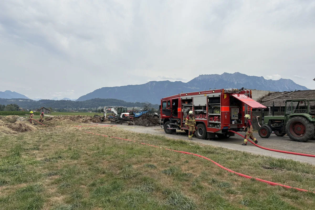 Ein rotes Feuerwehrfahrzeug steht auf einem Feld, während Feuerwehrleute Schläuche handhaben. Im Hintergrund sind Berge sichtbar.