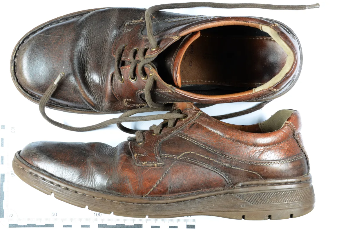 Ein Paar braune, abgenutzte Lederschuhe mit Schnürsenkeln liegt auf einem weißen Hintergrund. Maßstabsmarker sind sichtbar.
