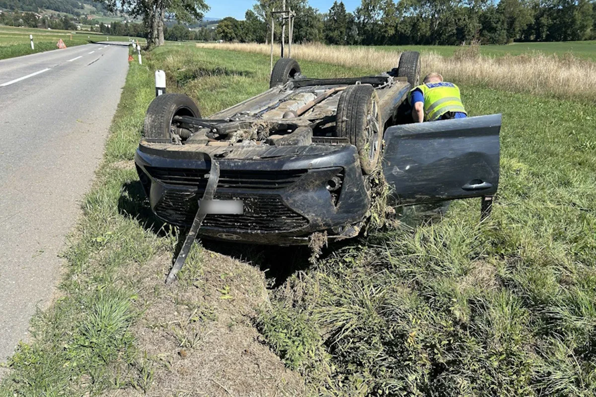 Ein Auto liegt auf dem Dach im Gras neben einer Landstraße. Ein Polizist untersucht das Fahrzeug.