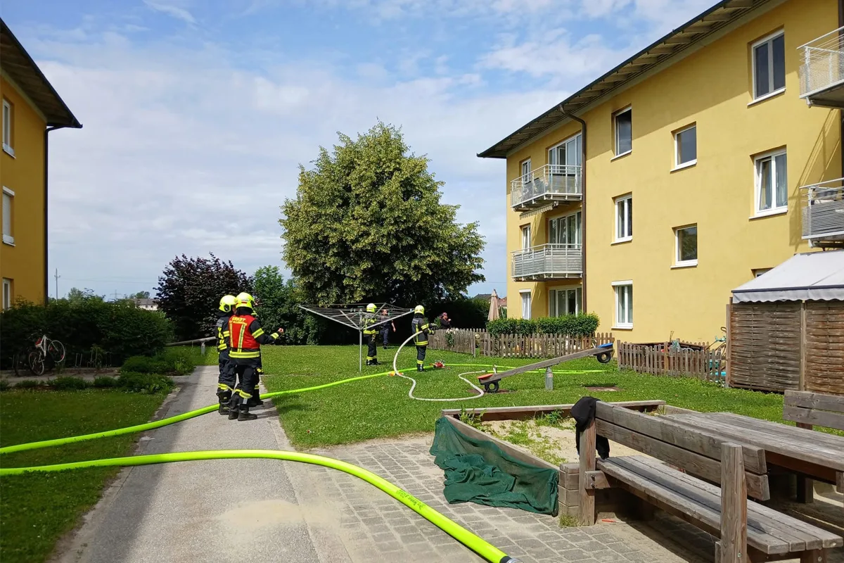 Mehrere Feuerwehrleute in Schutzkleidung stehen auf einem Rasen vor einem gelben Wohnhaus. Sie tragen Schläuche.