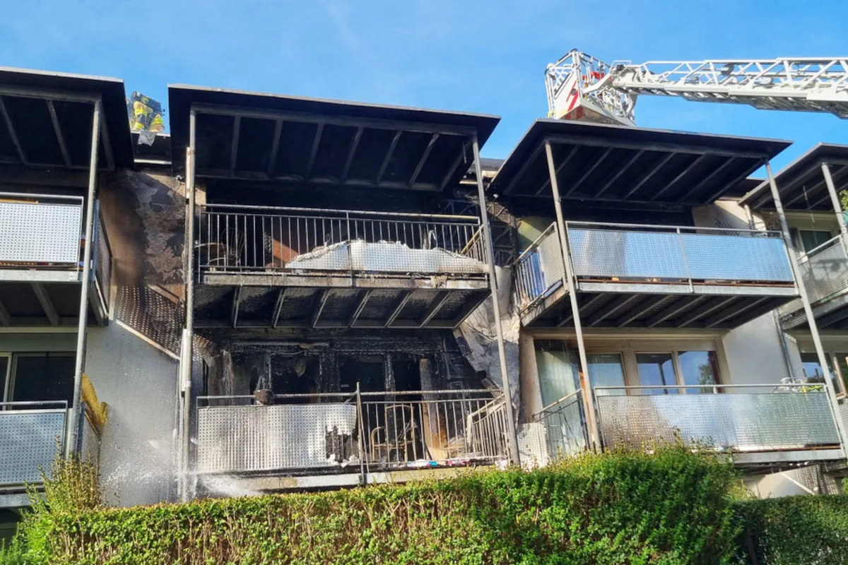 Ein Wohnhaus mit erheblichen Brandschäden an einem Balkon, von der Feuerwehr bearbeitet, ist zu sehen. Die Fassade ist verrußt und eine Drehleiter ist im Einsatz.