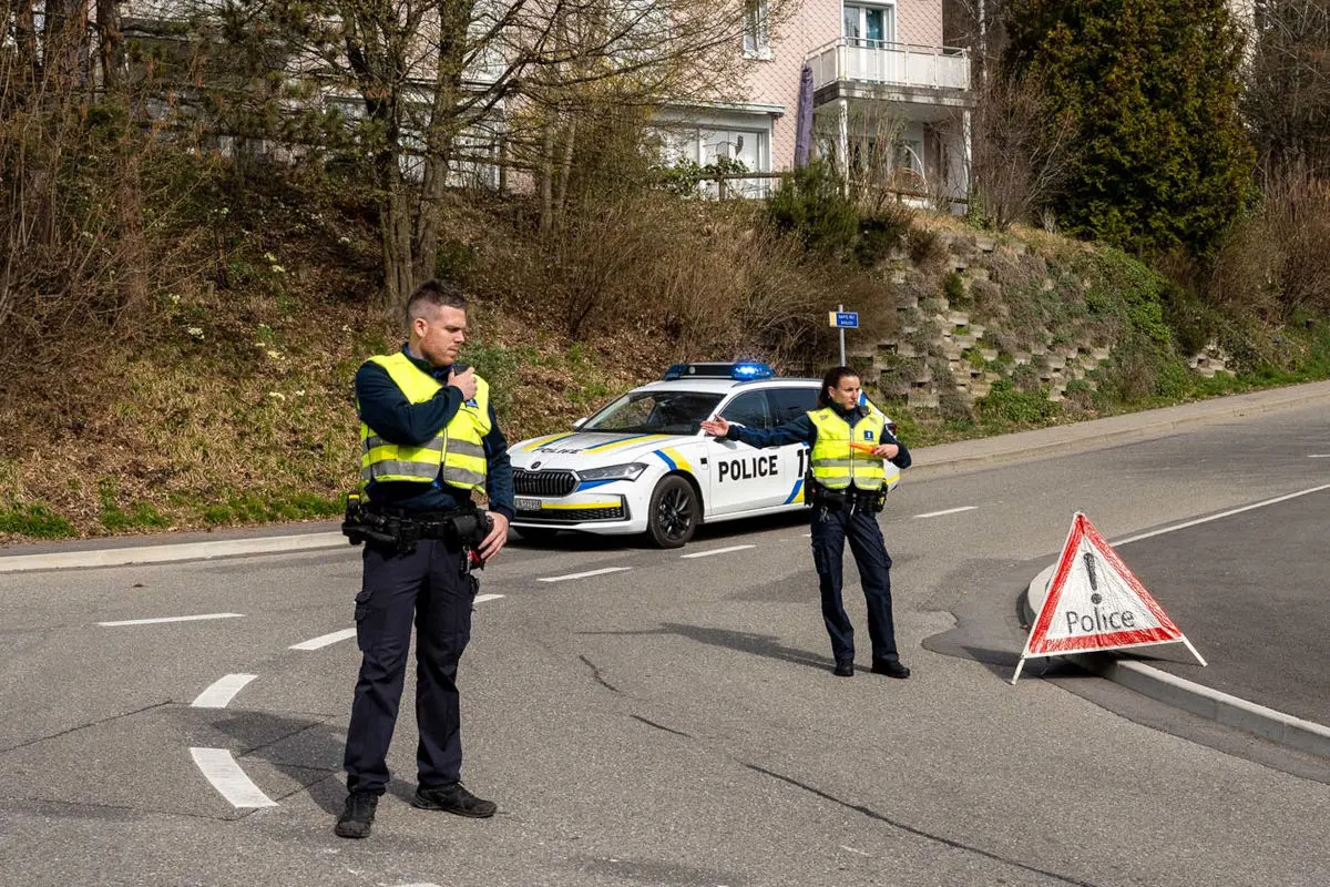 Zwei Polizisten in Warnwesten sichern eine Straße mit einem Polizeiauto und einem Warnschild. Im Hintergrund sind Wohnhäuser sichtbar.