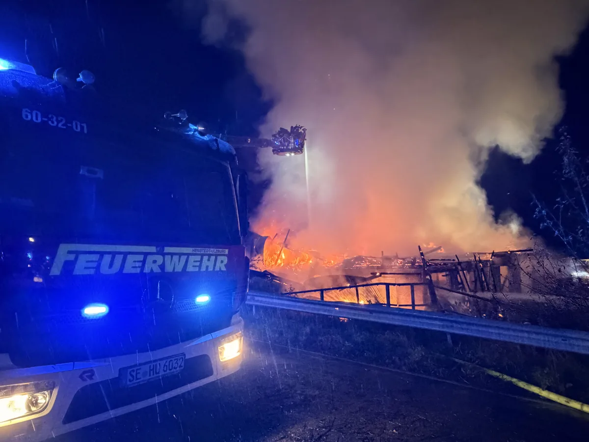 Ein Feuerwehrfahrzeug steht in der Nacht vor einem brennenden Gebäude mit starker Rauchentwicklung. Blaulicht ist zu sehen.