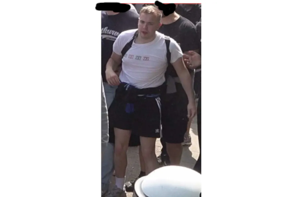 Ein Mann in einem weißen T-Shirt und schwarzen Shorts wird von mehreren Personen begleitet. Er scheint in Bewegung zu sein.