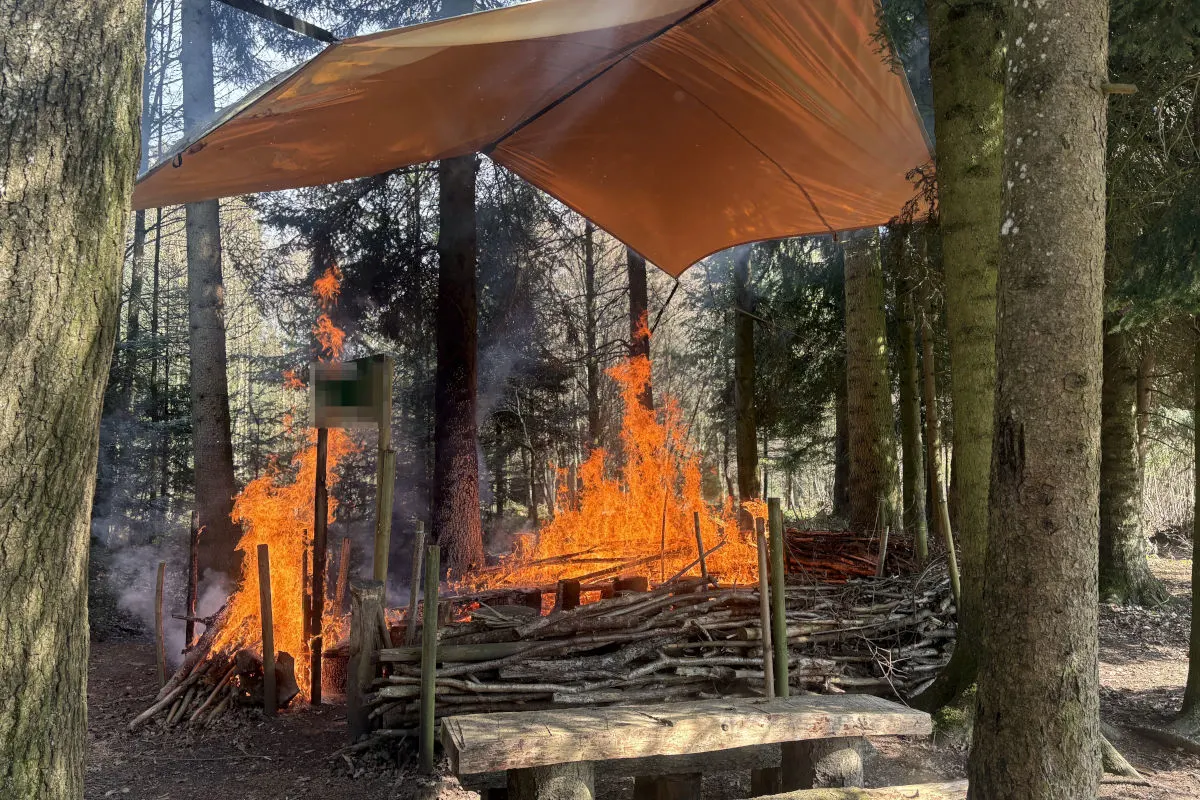 Ein Lagerfeuer brennt unter einer Plane in einem Wald, umgeben von hohen Bäumen und Holzwänden.