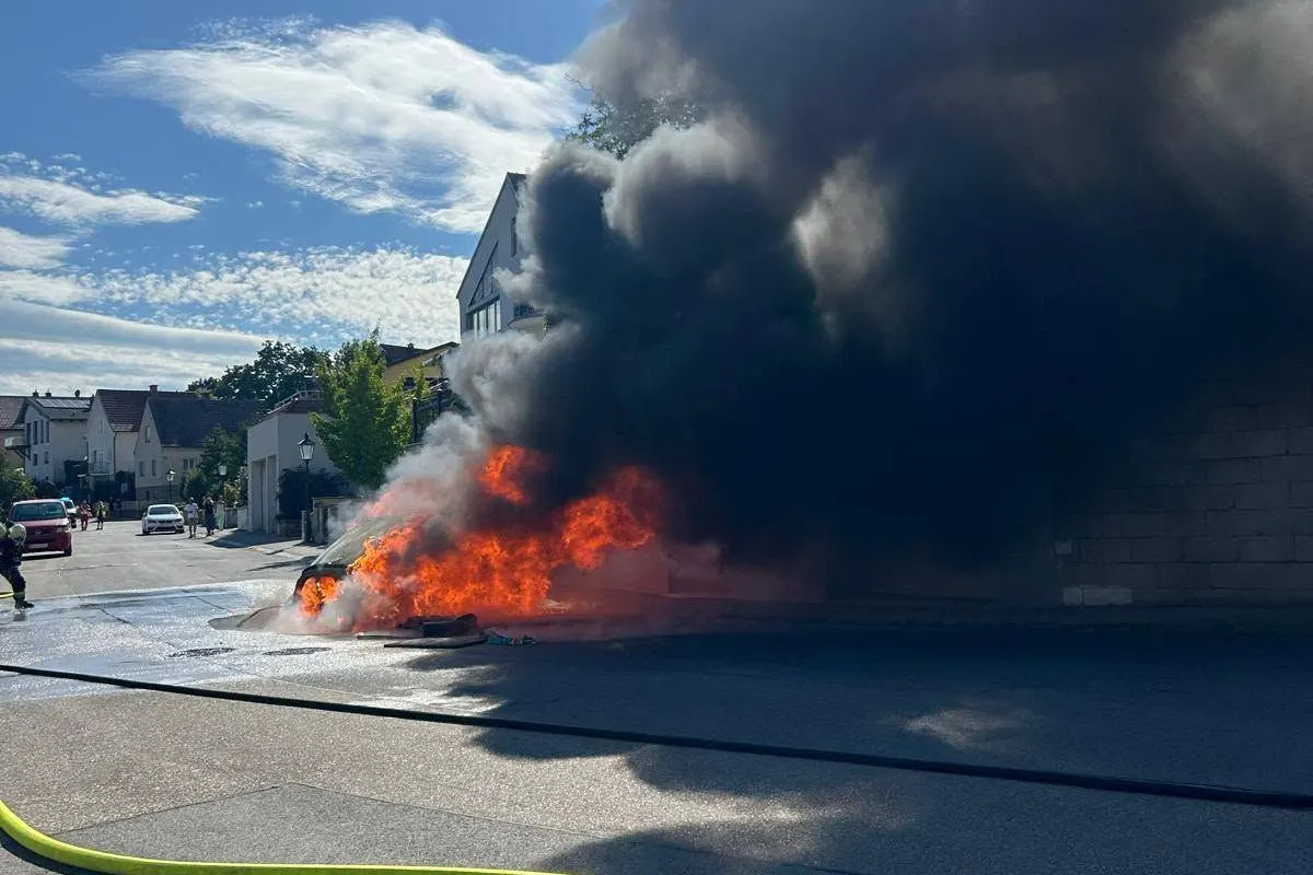 Ein Auto steht auf einer Wohnstraße in Flammen, dicke schwarze Rauchwolken steigen auf. Im Hintergrund sind Häuser sichtbar.