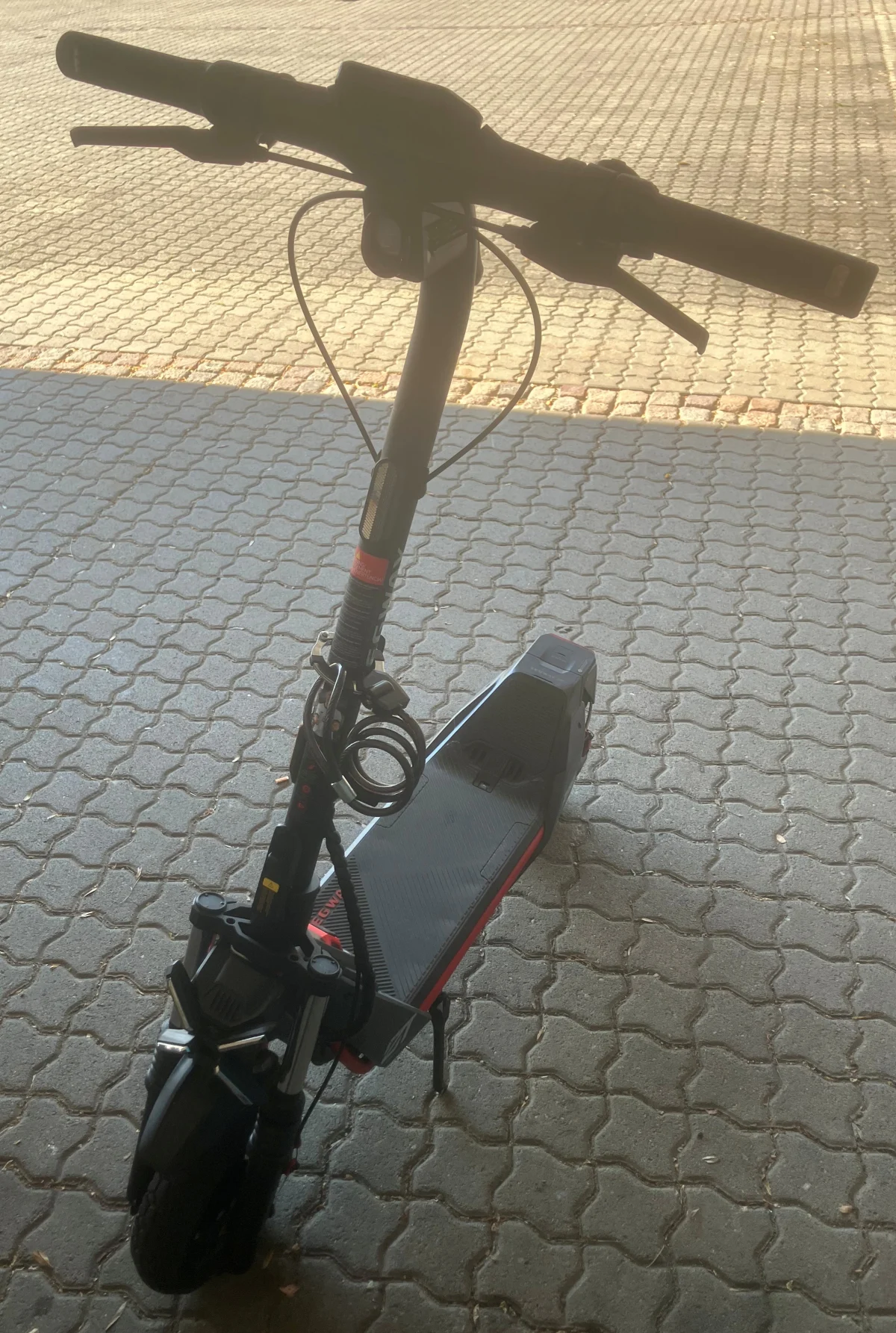 Ein schwarzer E-Scooter steht auf einem gepflasterten Gehweg im Freien. Er ist unbenutzt und mit einem Schloss gesichert.