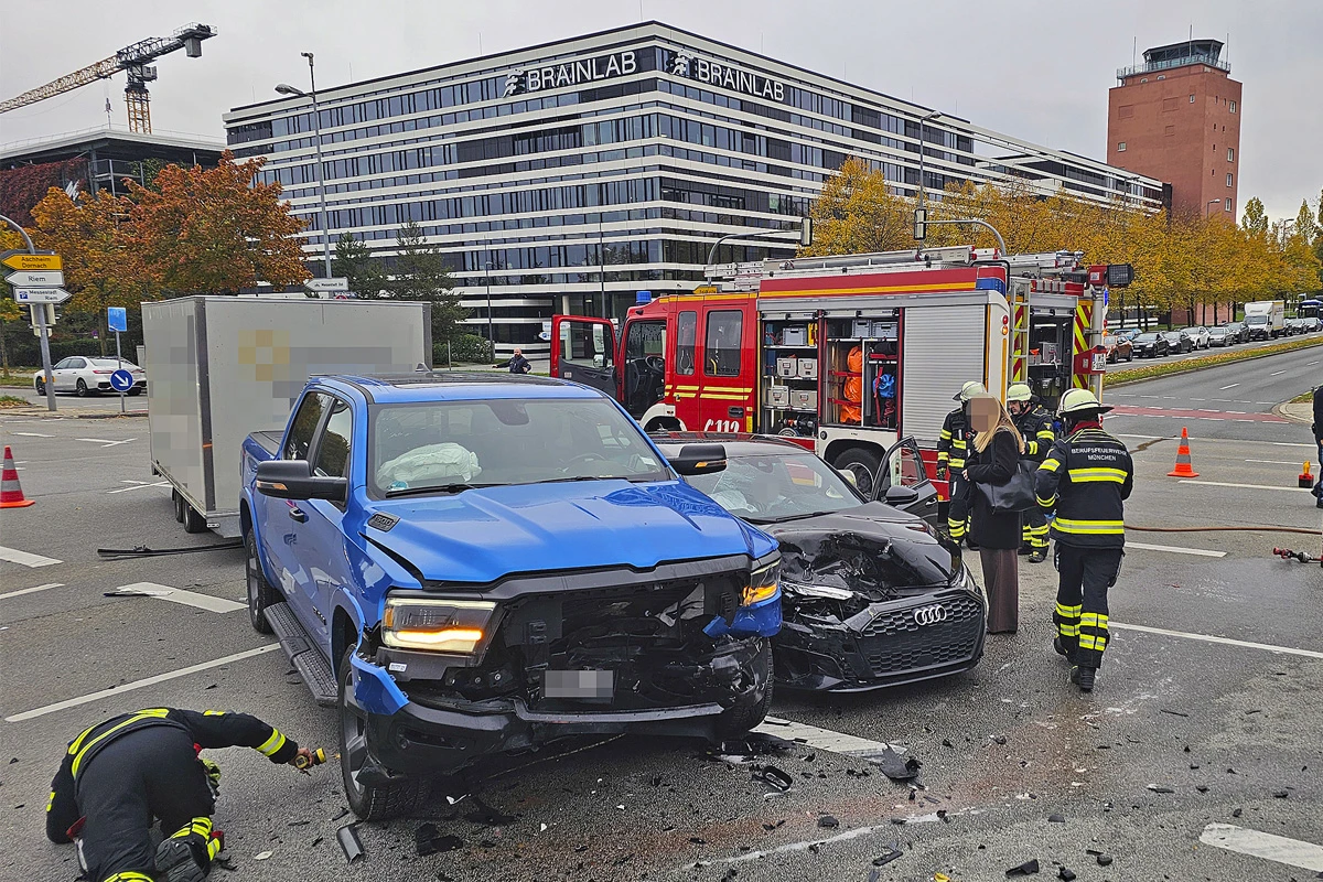 Ein blauer Pickup und ein schwarzer Audi mit deutlichen Schäden stehen nach einem Unfall vor einem modernen Bürogebäude. Feuerwehrleute sind anwesend.