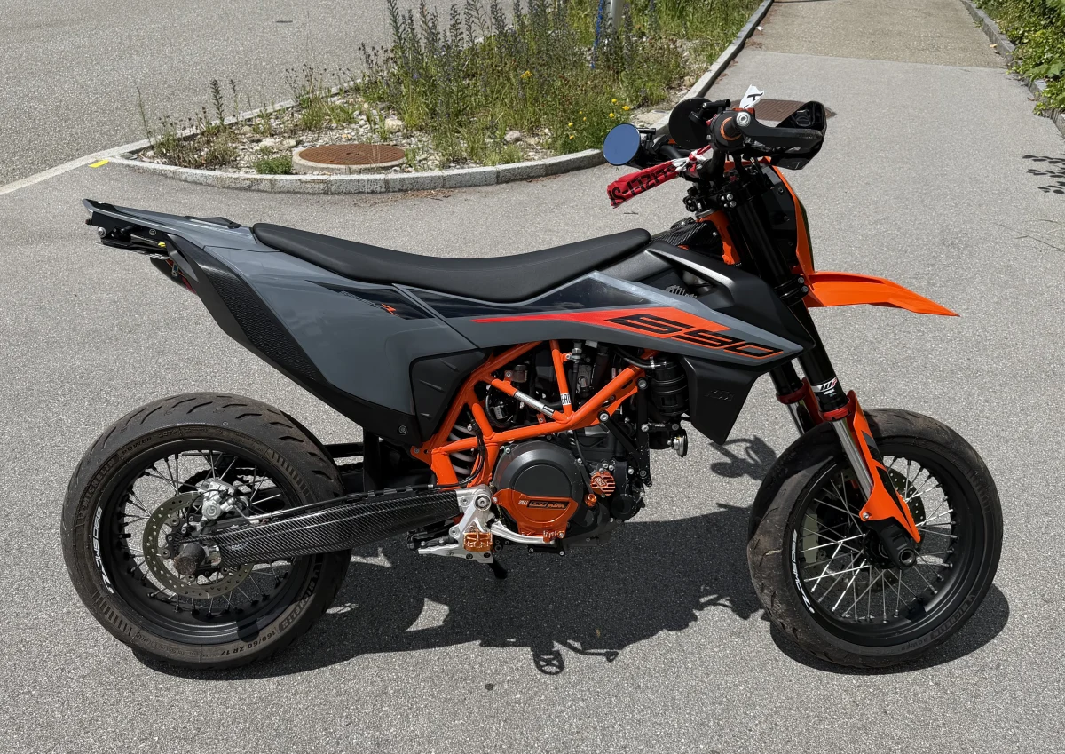 Ein orange-schwarzes Motorrad steht auf einem leeren Parkplatz neben einer Bordsteinkante mit Unkrautbewuchs.