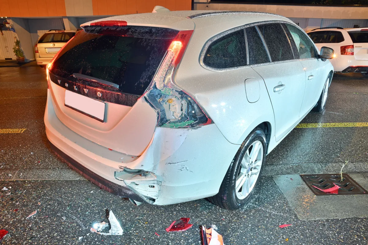 Das Bild zeigt einen weißen Volvo-Kombi mit beschädigter Rückleuchte und Kratzern am Heck, aufgenommen nachts auf einem nassen Parkplatz.