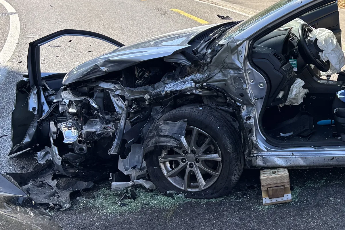 Ein stark beschädigtes silbernes Auto steht mit offenem Airbag und offenem Tür auf einer Straße nach einem Unfall. Glassplitter sind auf dem Boden verstreut.