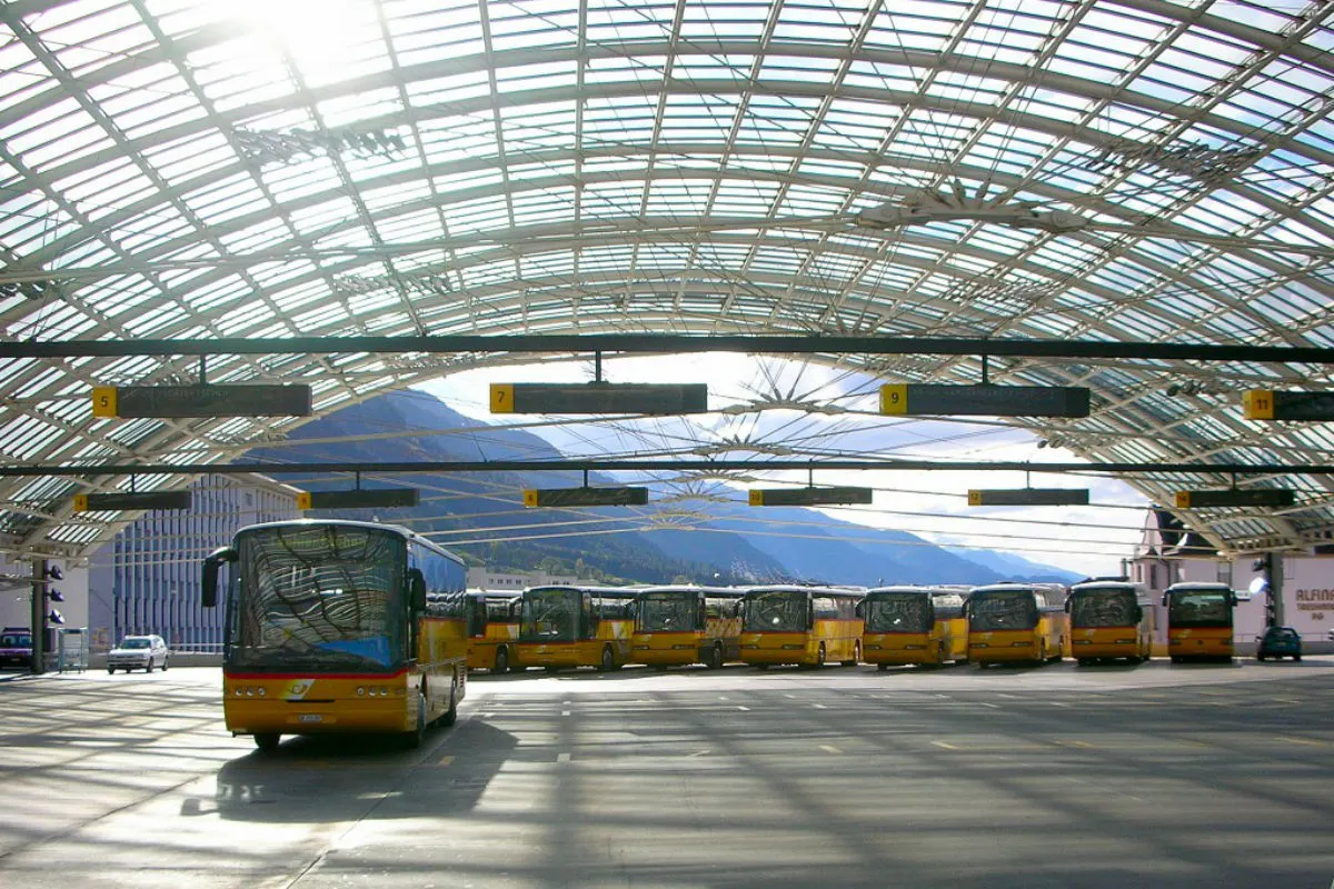 Im Vordergrund steht ein gelber Bus in einem überdachten Busbahnhof. Im Hintergrund sind weitere geparkte Busse und Berge sichtbar.