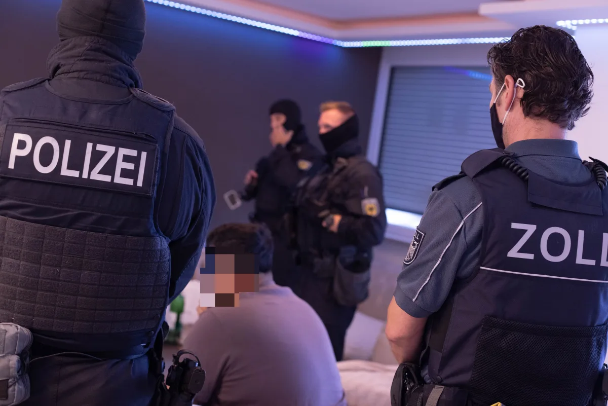 Mehrere Polizisten und Zollbeamte stehen in einer Wohnung, ein verdächtiger Mann sitzt im Vordergrund. Die Beamten tragen Schutzwesten mit Kennzeichnungen.
