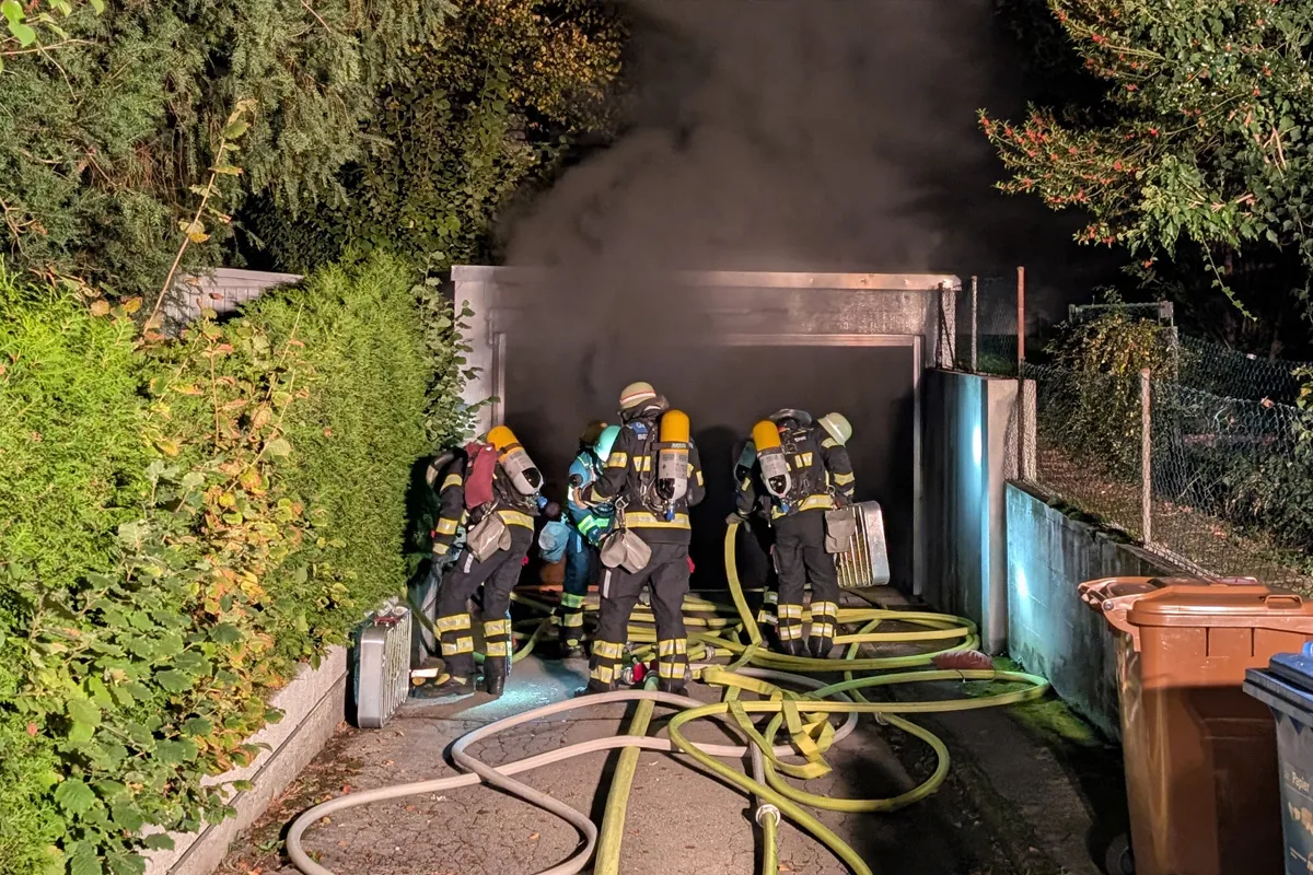Mehrere Feuerwehrleute mit Atemschutzgeräten stehen vor einer stark verrauchten Garage. Sie sind von Schläuchen umgeben.