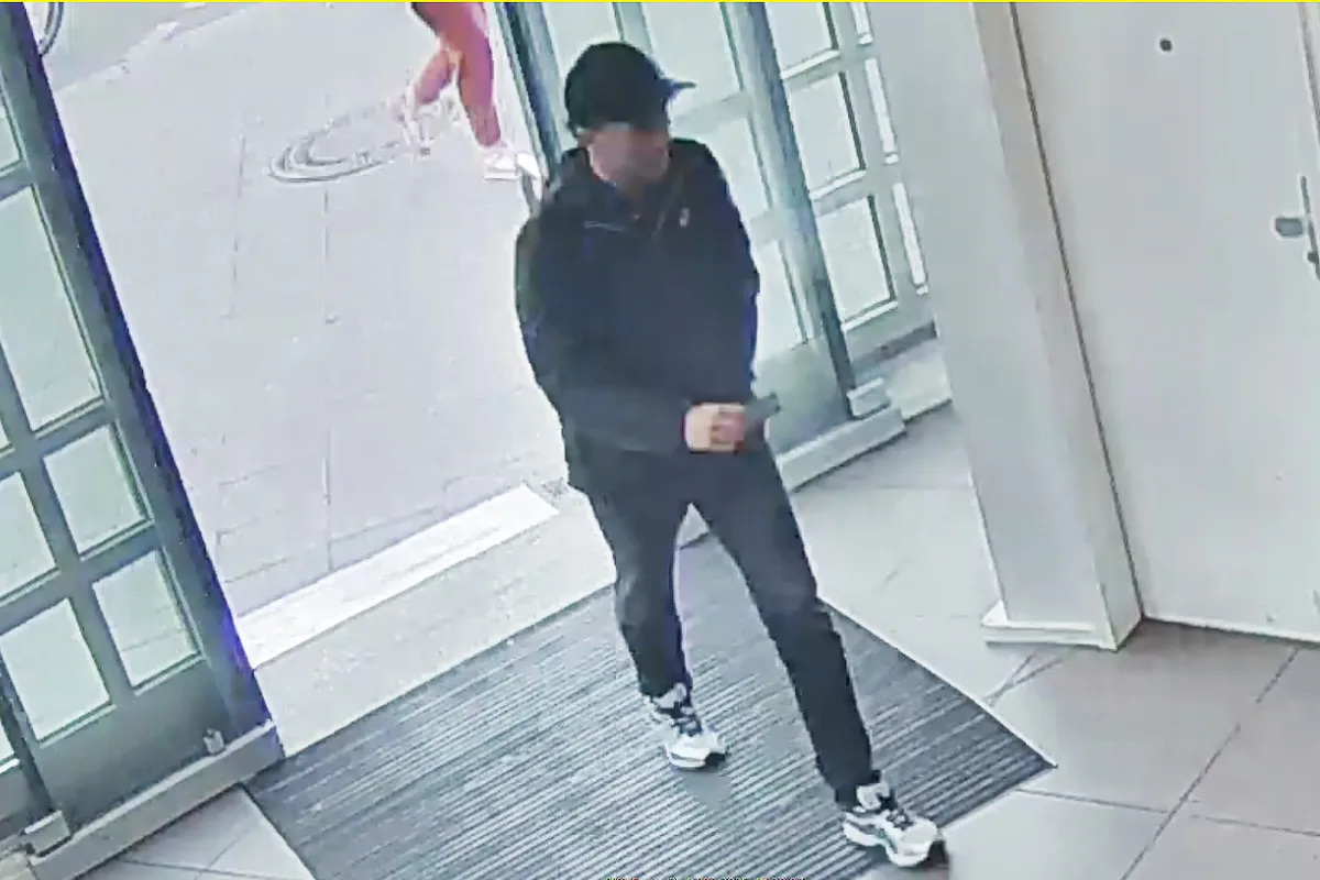 Eine Person in dunkler Kleidung und Mütze betritt einen Gebäudeeingang auf einer Überwachungskameraaufnahme. Im Hintergrund ist der Bürgersteig zu sehen.