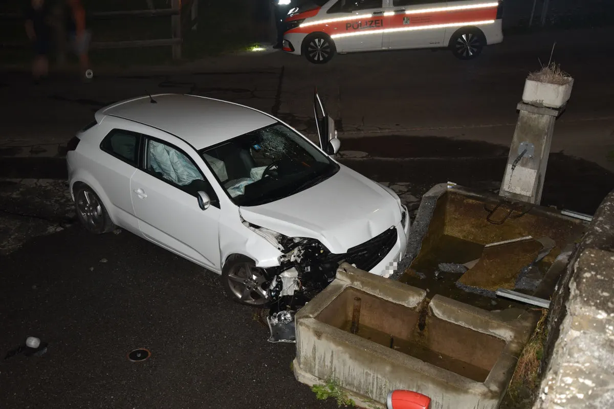 Ein weißes Auto mit stark beschädigter Front steht neben einem Betonsockel. Im Hintergrund ist ein Polizeifahrzeug sichtbar.