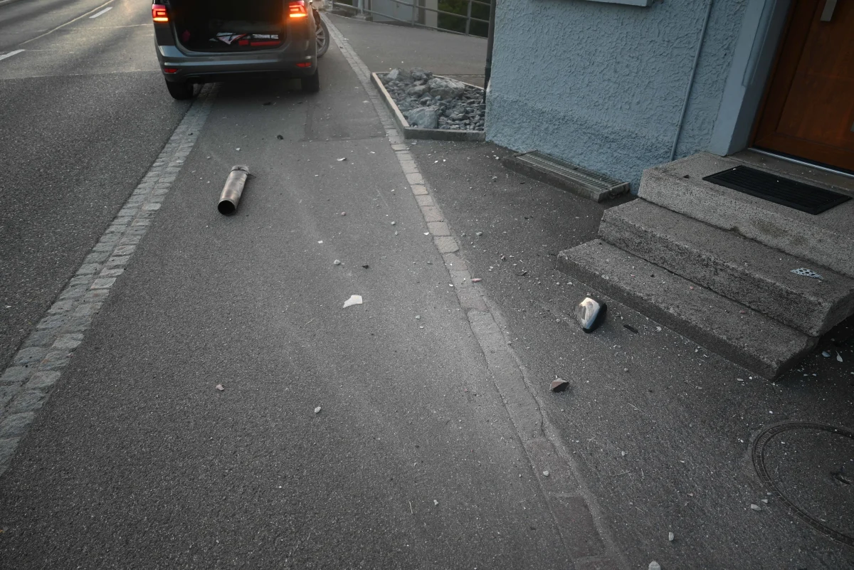 Ein Auto steht mit offenem Kofferraum auf einer Straße. Auf dem Gehweg sind Trümmer verteilt, nahe einer Haustreppe.