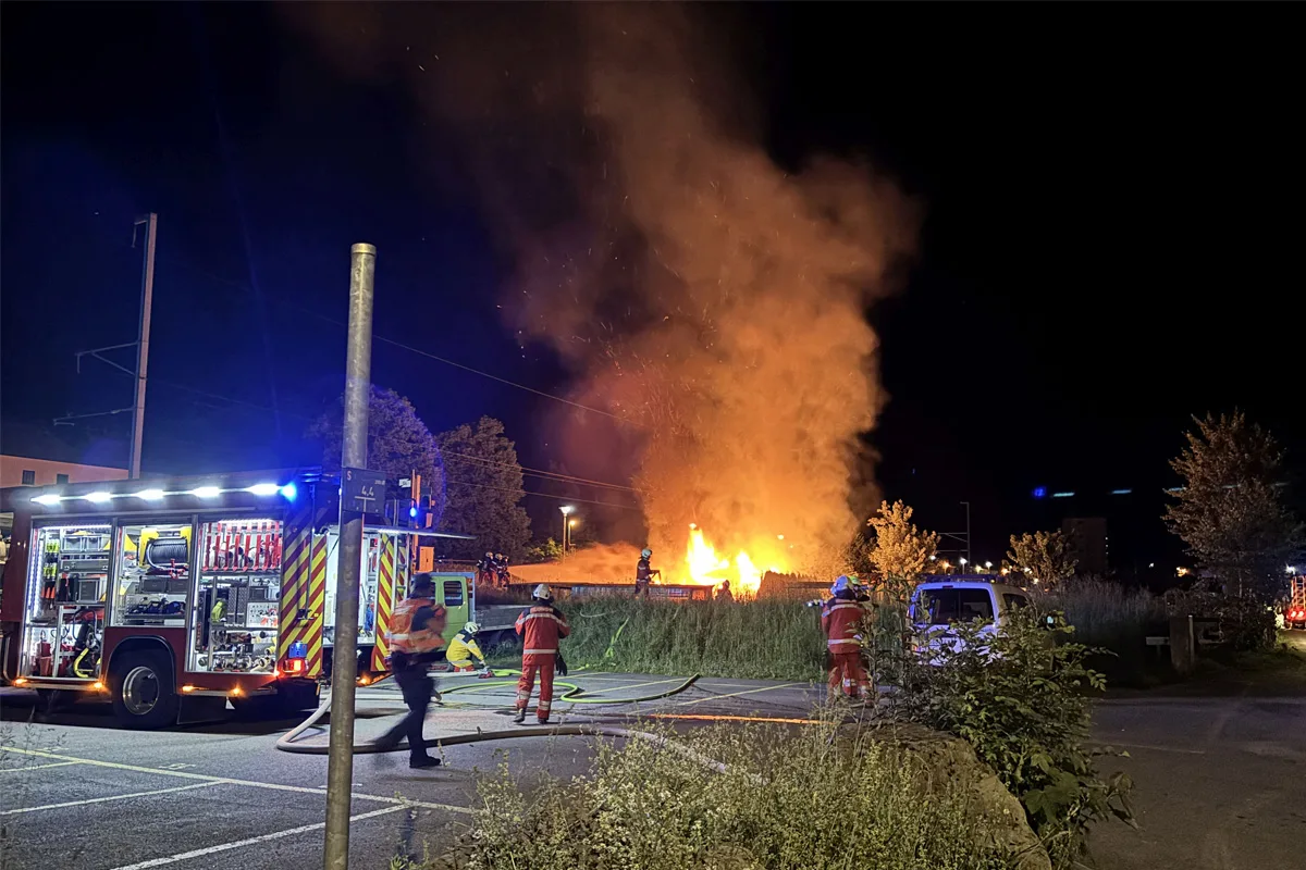 Bei Nacht bekämpfen Feuerwehrleute einen großen Brand auf einem offenen Gelände. Ein Löschfahrzeug ist vor Ort aktiv.