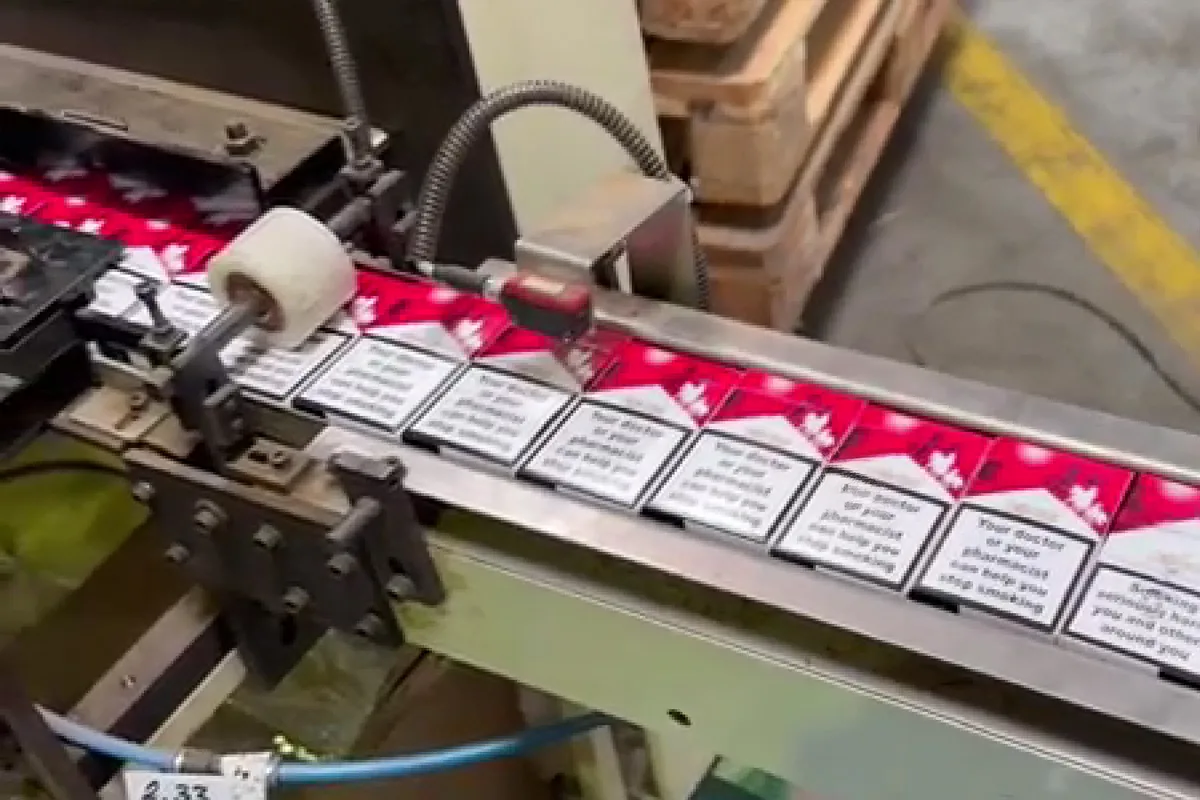 Ein Förderband transportiert Zigarettenpackungen in einer Produktionsumgebung. Maschinen und Verpackungen sind sichtbar.