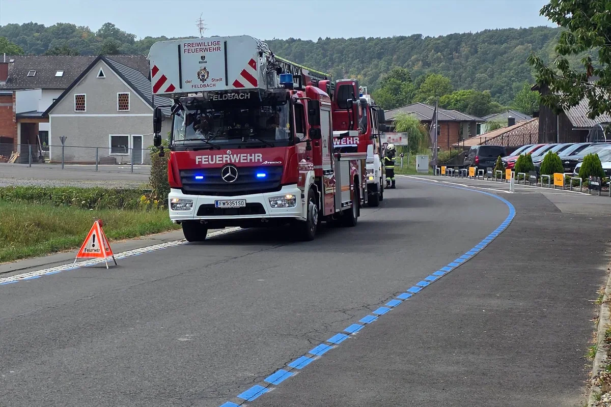 Ein rotes Feuerwehrfahrzeug mit der Aufschrift 'Feuerwehr' und Blaulicht fährt auf einer Landstraße. Im Hintergrund sind Häuser und Bäume zu sehen.