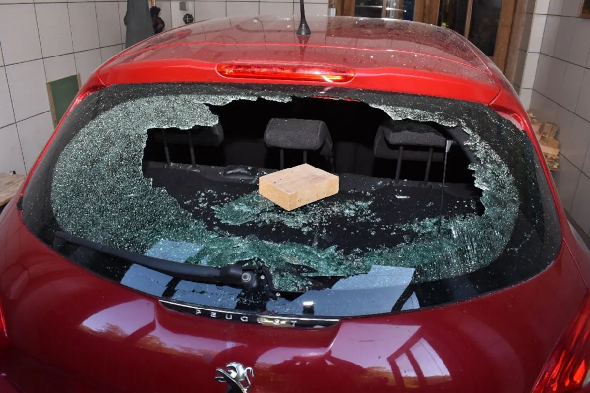 Das Bild zeigt die zerbrochene Heckscheibe eines roten Autos, durch die ein Backstein geworfen wurde. Der Backstein liegt sichtbar auf der Ablage im Inneren.