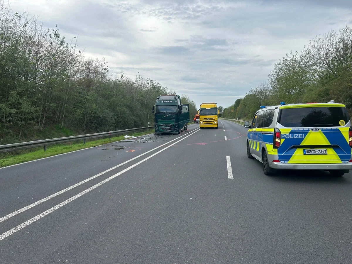 Ein beschädigter Lkw steht auf einer zweispurigen Landstraße, flankiert von einem Polizeiauto und einem Abschleppwagen. Auf der Straße sind Trümmer verteilt.